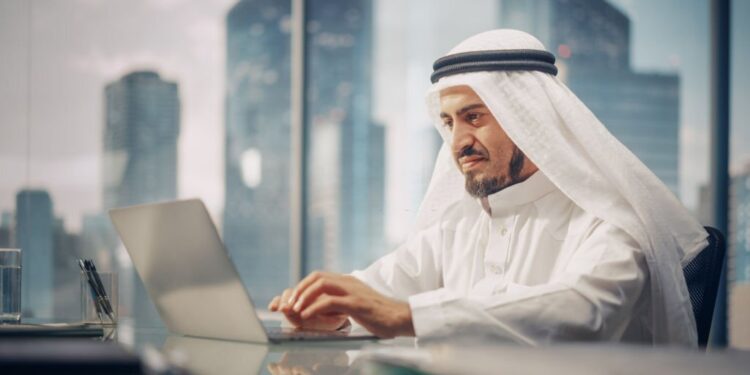 Successful Arab Businessman in White Traditional Outfit Sitting in Office and Working on Laptop Computer. Business Manager Planning Corporate Strategy. Saudi, Emirati, Arab Businessman Concept.
